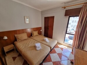 A hotel room with two beds, a wooden headboard, a small bedside table with a lamp, a window with curtains, and a view of the street outside. - Cerro Atlântico by Umbral (Albufeira)