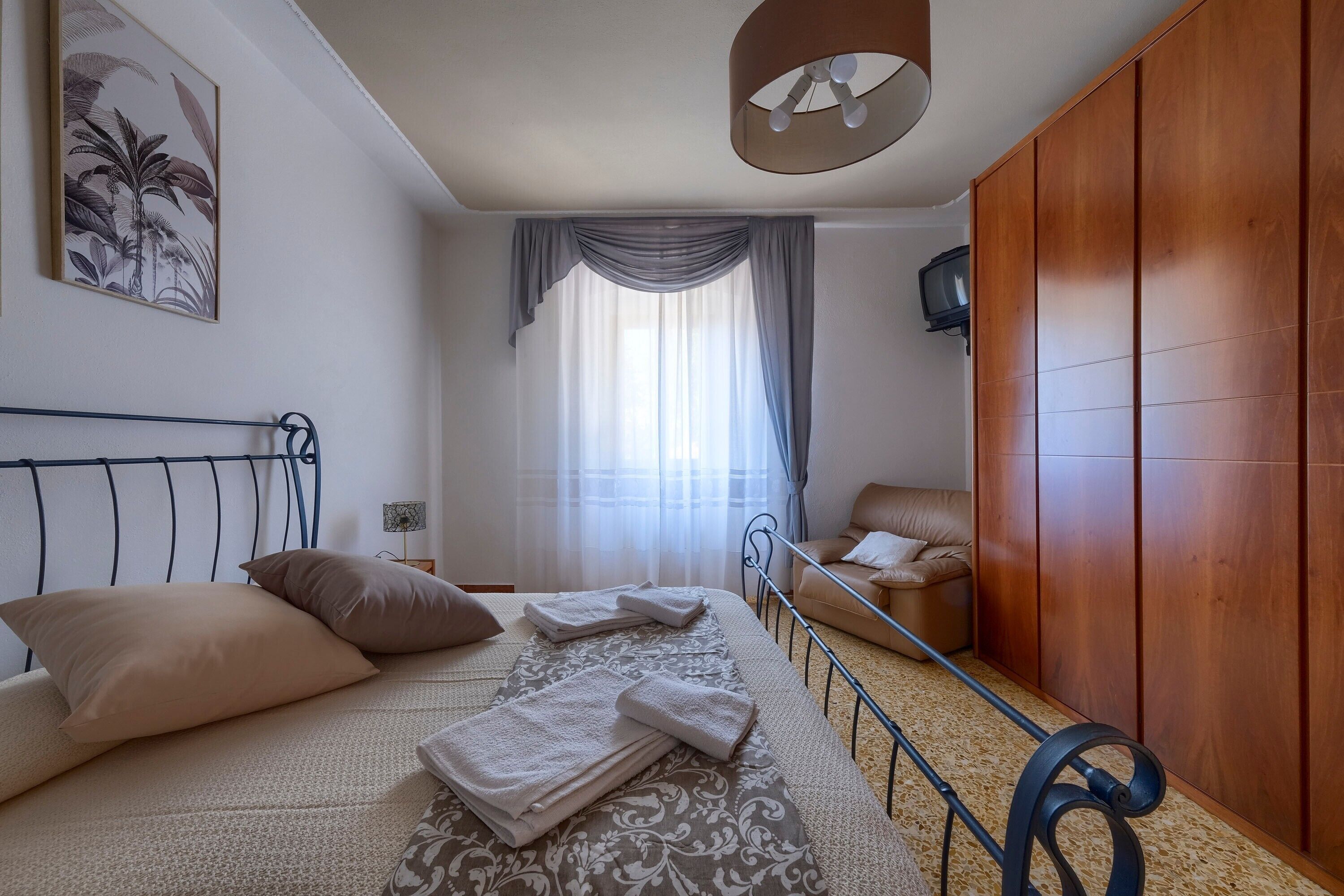 Un dormitorio con cama, almohadas, mesita de noche, armario de madera, televisor y ventana con cortinas.