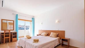 Un dormitorio con una cama grande, un escritorio y una silla. Hay una ventana con cortinas, una lámpara de pared y vistas a edificios.