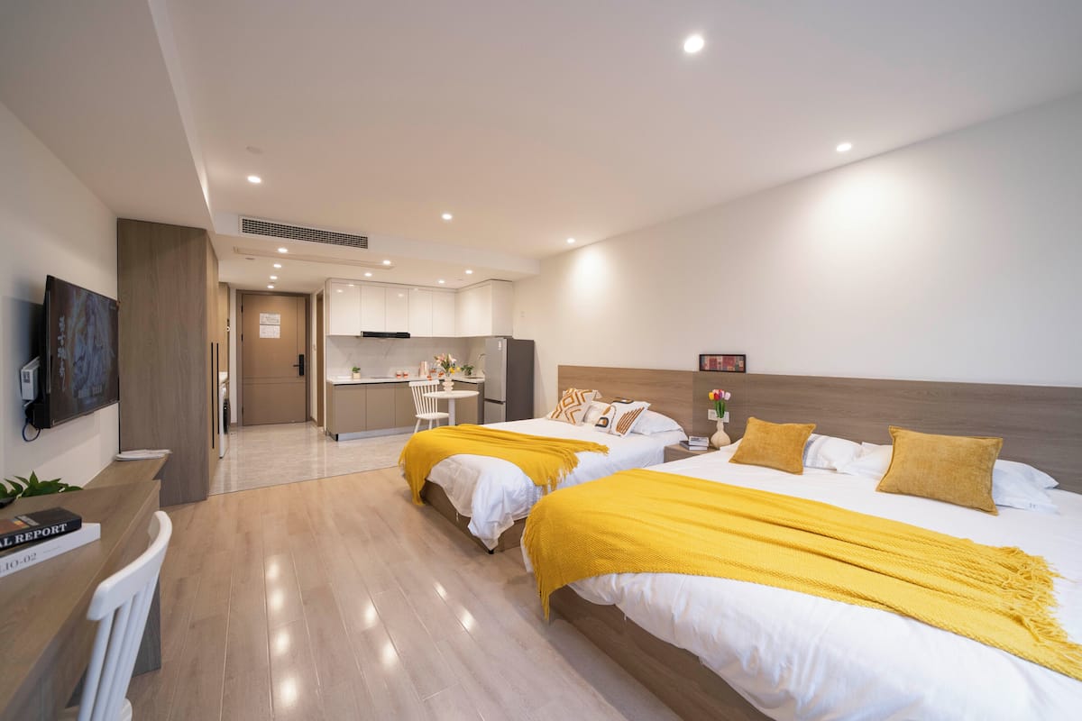 A modern hotel room with two beds, a desk, and a kitchenette.