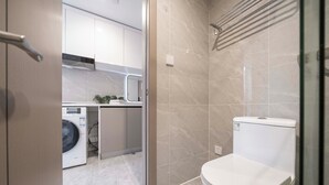 A modern bathroom with a glass shower enclosure, a toilet, and a washing machine in the adjacent room. - Book and Bed-QuYang Road (Shanghai)