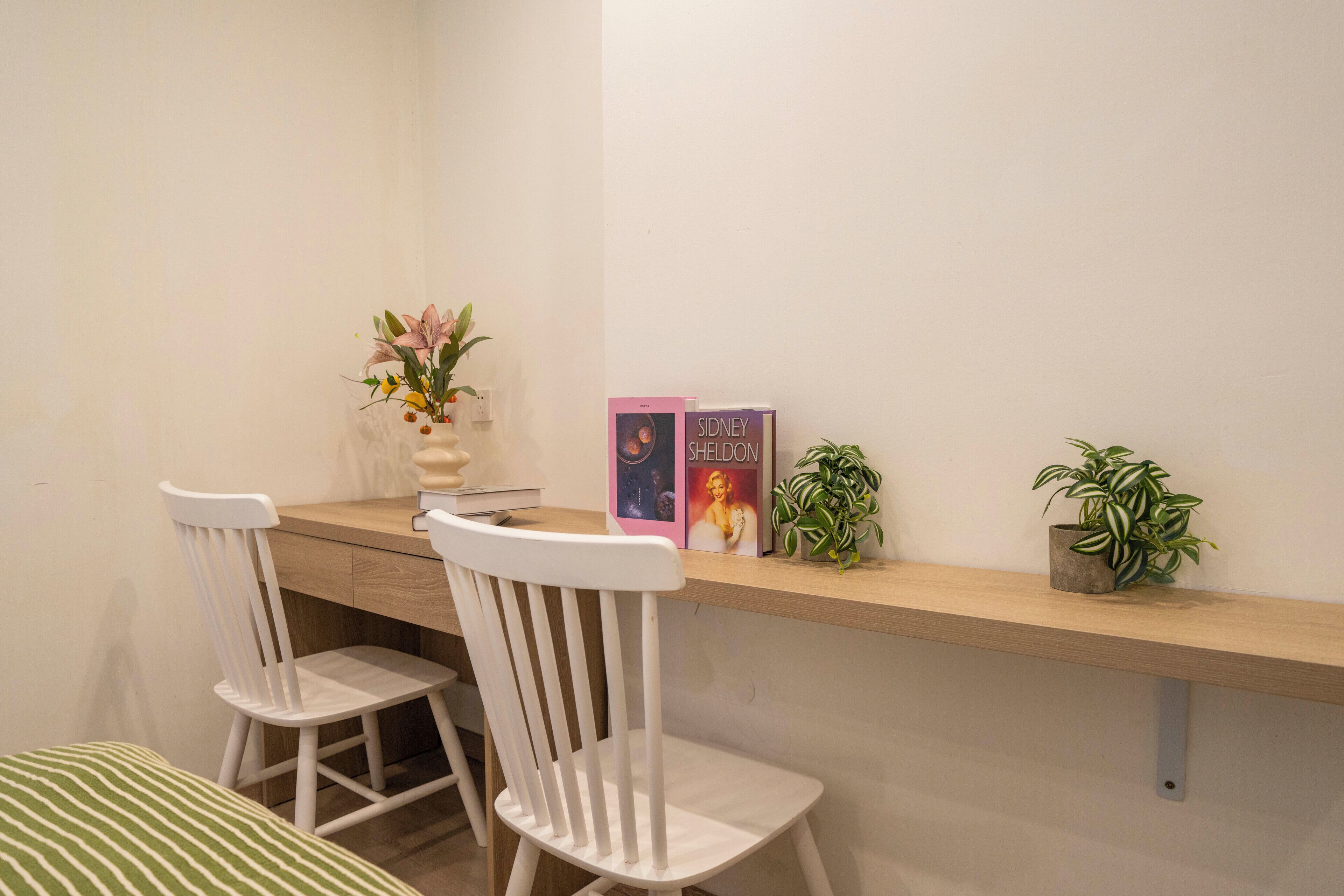 A small, neatly arranged workspace with two white chairs, a wooden desk, a vase with flowers, two books, and two potted plants.