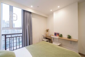 Une chambre d’hôtel moderne dotée d’un grand lit, d’un bureau avec deux chaises, d’une petite étagère avec des plantes et d’une fenêtre donnant sur des immeubles.