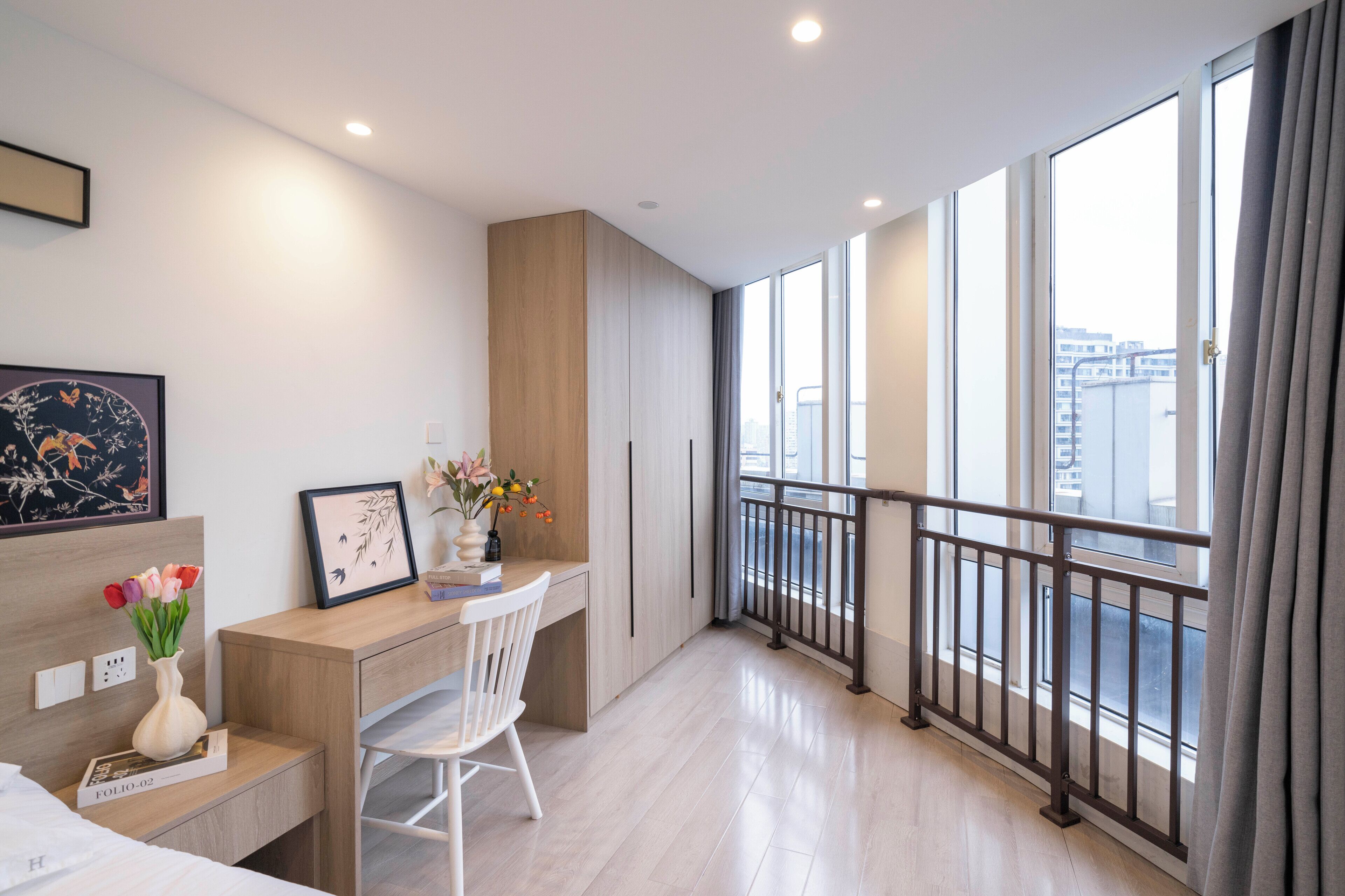 Una habitación de hotel moderna con un escritorio de madera, una silla blanca, ventanas amplias y un balcón con barandilla de metal.