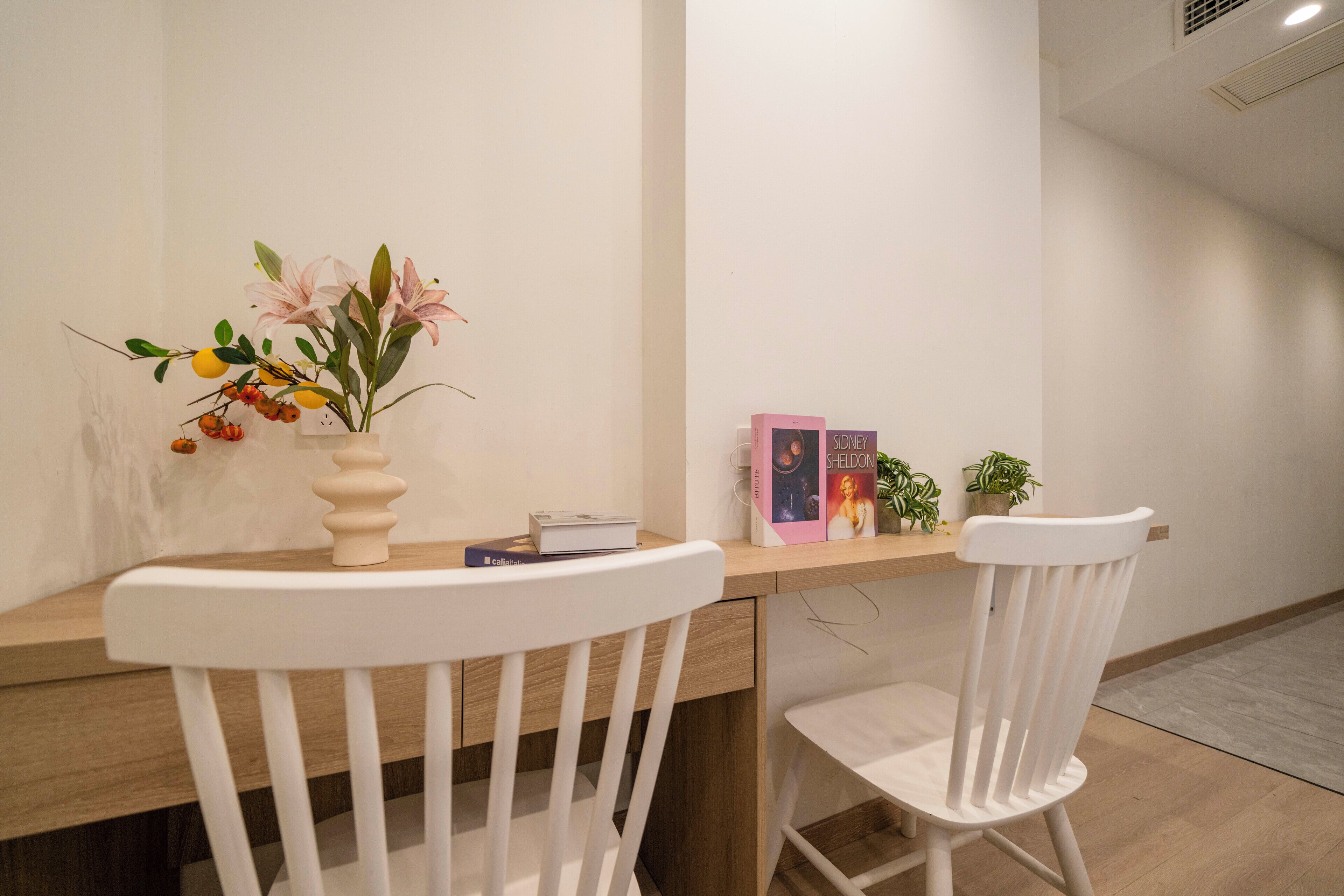 A modern room with a wooden desk and two white chairs. A vase with flowers and a book is on the desk.