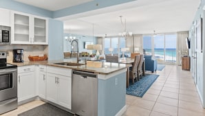 A modern kitchen with white cabinets, a dishwasher, and a coffee maker. There is a dining area with wicker furniture and a view of the ocean. - Great Beach Front Condo at Pelican Beach (Destin)