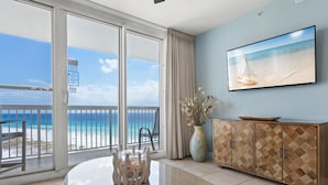 A room with a large sliding glass door leading to a balcony with a beach view, a round glass table with a vase, and a wooden cabinet with a painting of a sailboat. - Great Beach Front Condo at Pelican Beach (Destin)