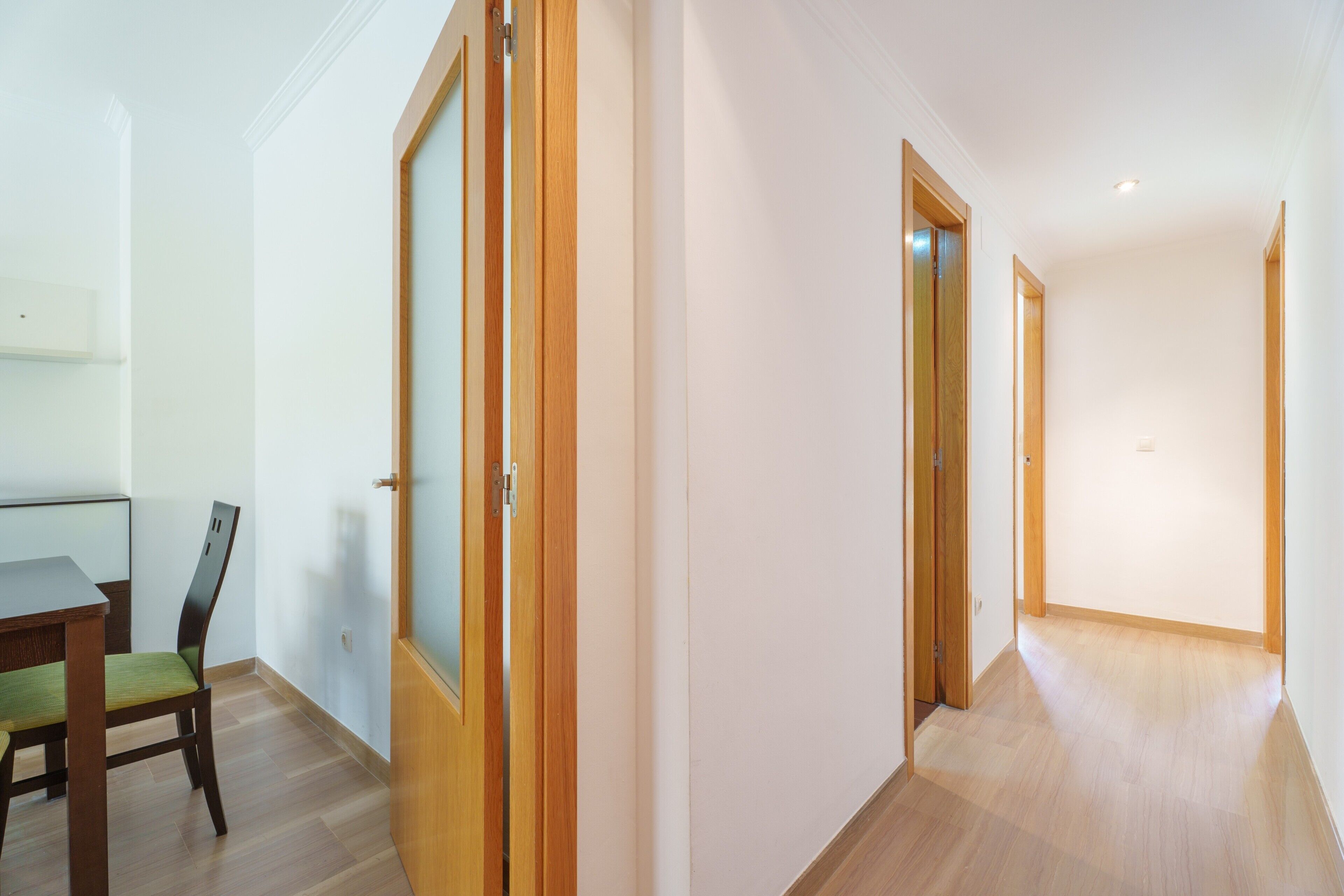 Un pasillo con suelo de madera, una zona de comedor con una mesa y sillas, y una puerta que da a otra habitación.