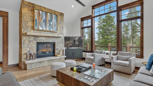A modern living room with a stone fireplace, a mounted TV, and large windows offering a view of trees. - Modern Luxury | Mountain Views | Hot Tub + Shuttle - Cherrybrook Chalet (Breckenridge)