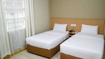 A hotel room with two single beds, a wooden headboard, a nightstand with a phone, and a window with sheer curtains.