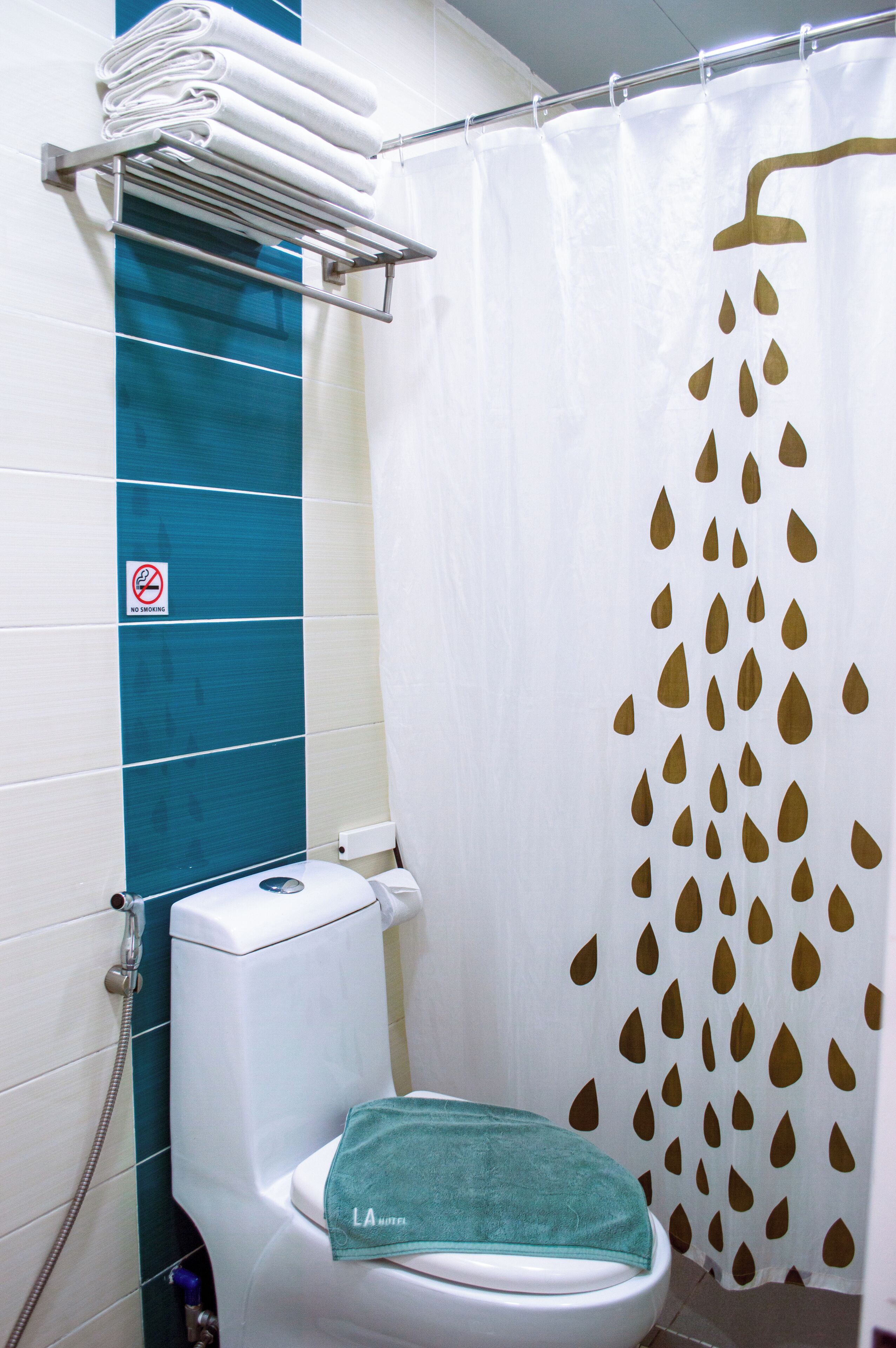 Un baño con inodoro blanco, cortina de ducha con diseño de gotas de agua, toallero con toallas dobladas y un cartel de 'No Smoking' en la pared.