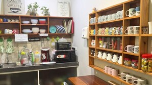 Ett litet café med ett träbord och stolar, hyllor med muggar och köksredskap, och en skylt som visar att det serveras kaffe.