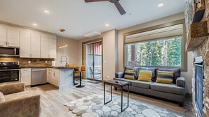 A modern kitchen with a central island, a dining area with a sofa and table, and a large window with a view of trees.