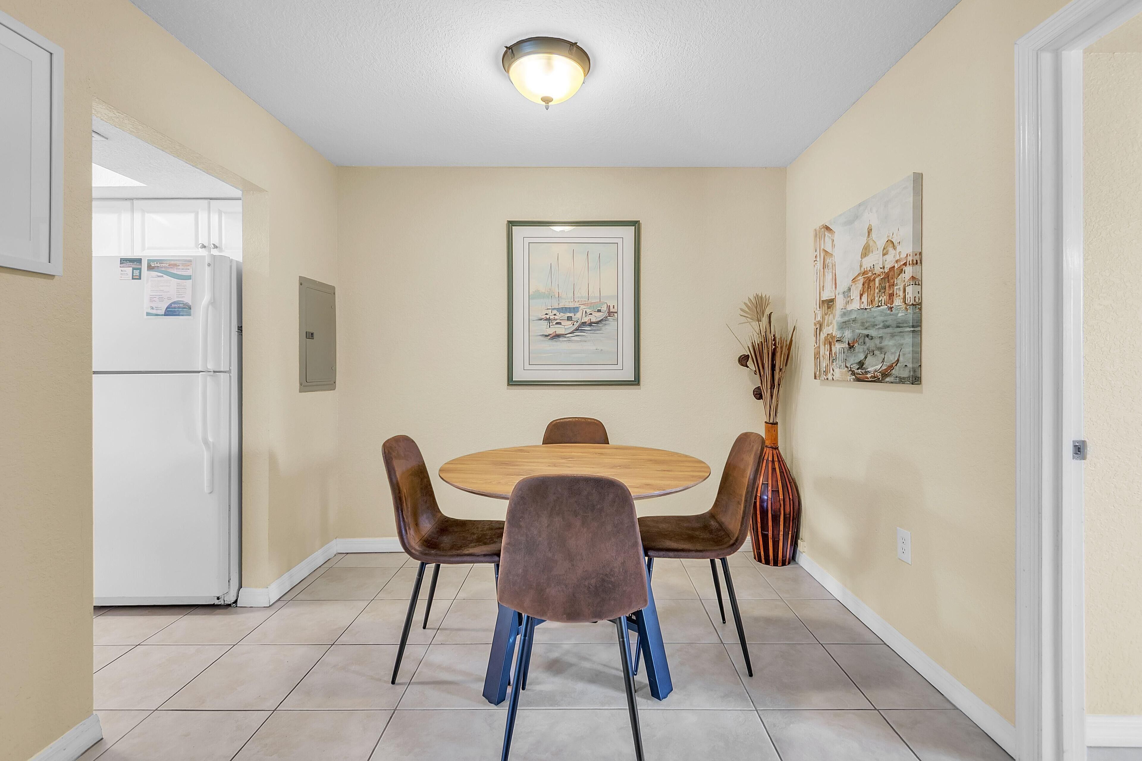 Un comedor con una mesa redonda, cuatro sillas, un refrigerador y dos cuadros colgados en la pared.