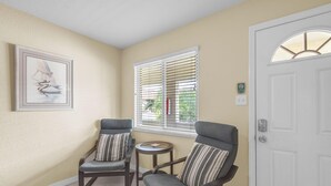 Une pièce comprenant deux fauteuils, une petite table ronde, une porte et une fenêtre avec des stores.