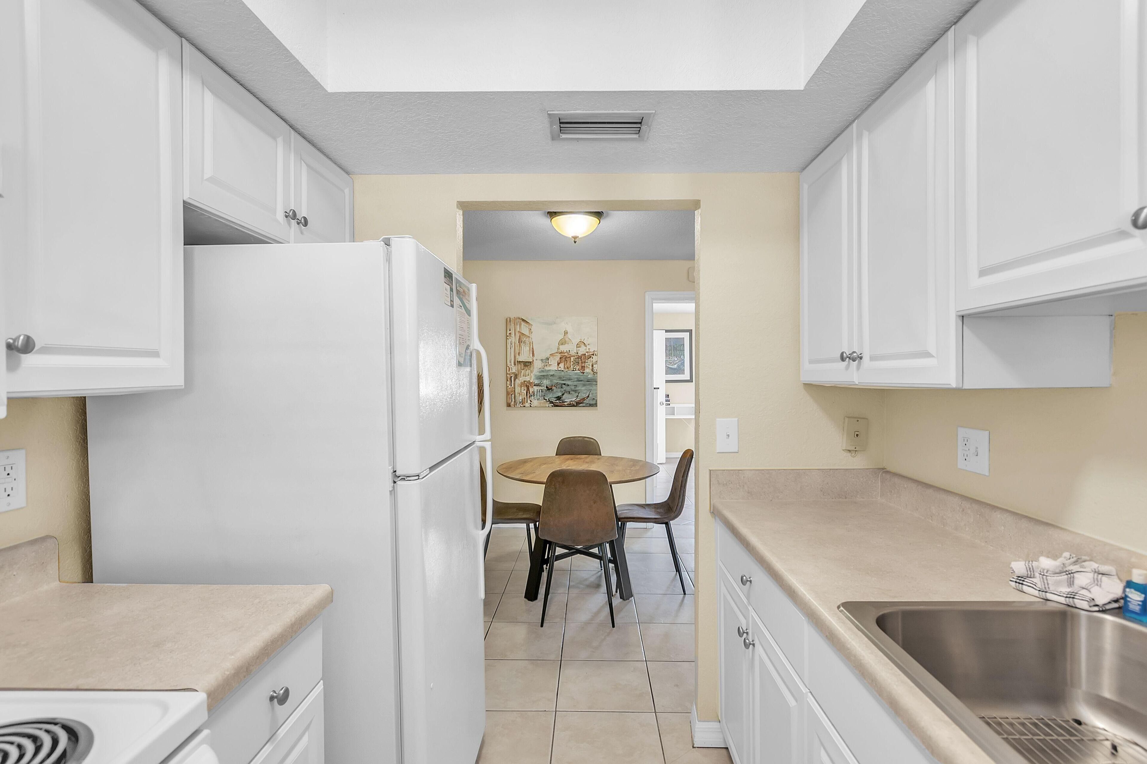 A compact kitchen with white cabinets, a refrigerator, a dining table with chairs, and a sink.