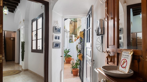 A hallway with a tiled floor, a wooden door, and a view into a room with a tiled floor and potted plants.