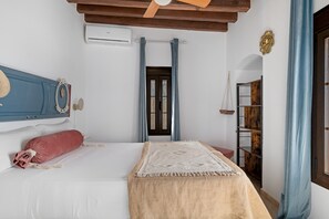A bedroom with a bed, a headboard, a window with curtains, a wooden shelf, and a door.