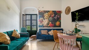 A living room with a sofa, chairs, a dining table, a TV, and a large floral painting.