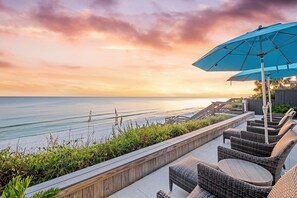 Une terrasse en bord de mer, meublée de mobilier en osier et agrémentée d’un parasol bleu, d’où l’on a une vue sur l’océan.