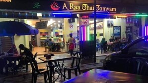 Een terras van een restaurant met tafels en stoelen, een neonbord boven de ingang en een auto in de buurt.