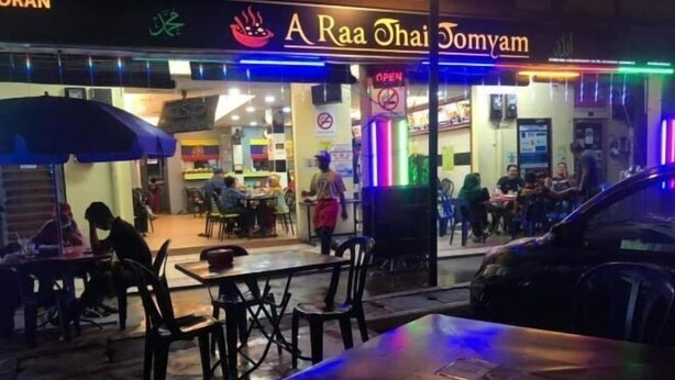 Egy étterem szabadtéri étkezőtere asztalokkal és székekkel, a bejárat felett egy neonfelirattal, és a közelben parkoló autóval.