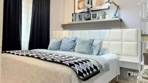Een slaapkamer met een bed, een nachtkastje, een lamp, een plank met boeken en decoratieve objecten, en een raam met gordijnen.