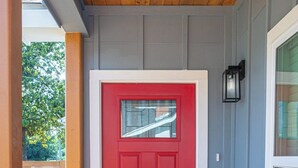 Una puerta roja con un ventanal pequeño, un buzón negro y una planta en maceta sobre una terraza de madera.