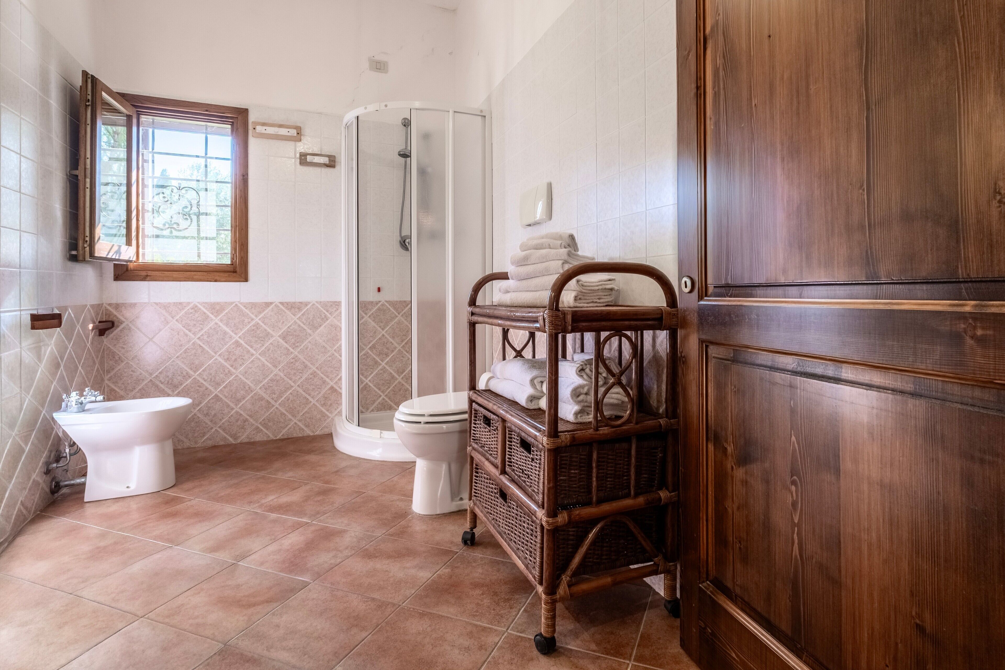 A bathroom with a shower, toilet, bidet, and a wicker shelf with towels.