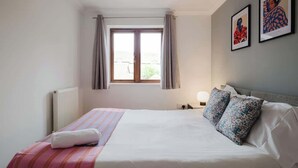 Une chambre d’hôtel avec un lit, une fenêtre avec des rideaux, un radiateur et deux tableaux encadrés accrochés au mur.