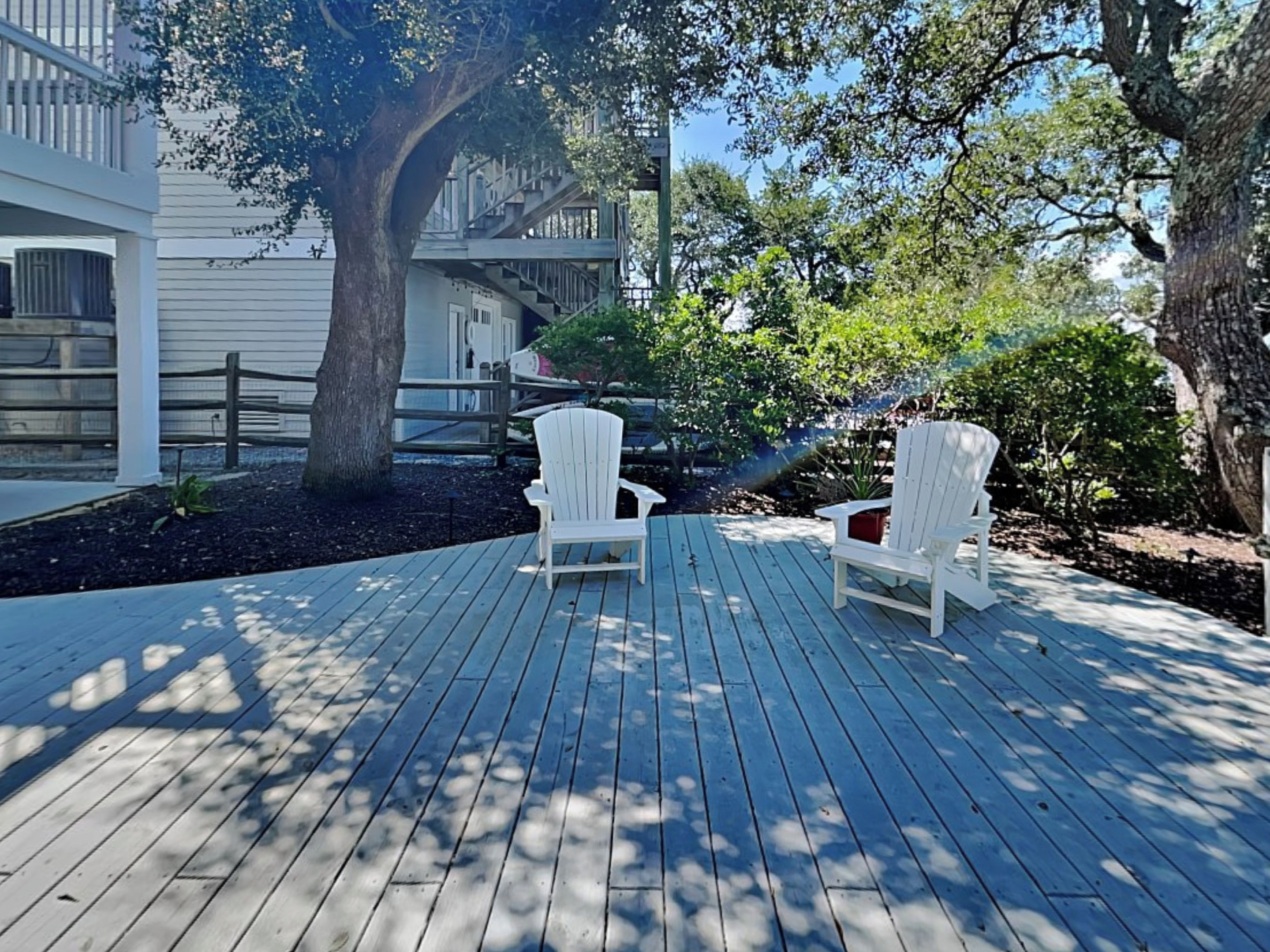 A wooden deck with two white Adirondack chairs, a large tree, and a building with a balcony in the background.