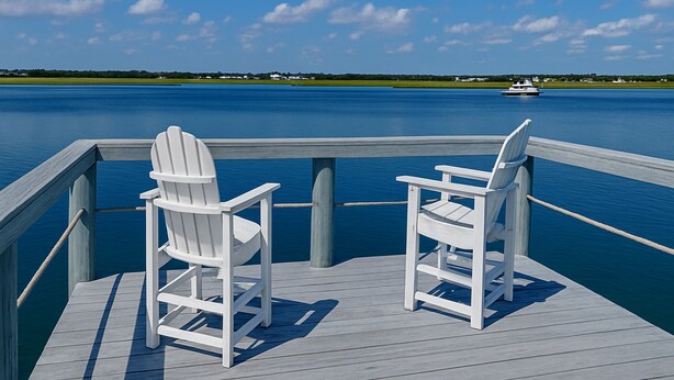 Een houten terras met twee witte Adirondack-stoelen gericht op een rustig meer, met een boot in de verte en huizen aan de horizon.