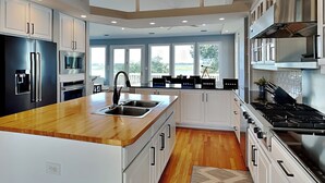 A modern kitchen with white cabinetry, a wooden countertop, stainless steel appliances, and large windows offering a view of the outdoors. - Shady Beaches by Topsail Realty (Holly Ridge)