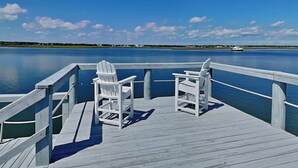 Een houten steiger met twee witte Adirondack-stoelen gericht op een rustig wateroppervlak. Een boot is in de verte te zien.