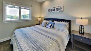 A bedroom with a large bed, two bedside tables, a window, and a wall sign that reads "NO WAKE ZONE". - Shady Beaches by Topsail Realty (Holly Ridge)