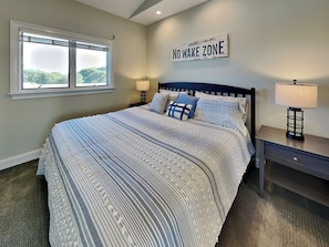A bedroom with a large bed, two bedside tables, a window, and a wall sign that reads "NO WAKE ZONE". - Shady Beaches by Topsail Realty (Holly Ridge)