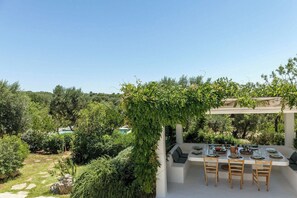 Ein Essbereich im Freien mit einer Pergola, einem Tisch für vier Personen und Blick auf die umliegende Bepflanzung und Hügel.