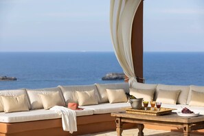 A seaside lounge with a cushioned seating area, a wooden table with a tray of drinks and grapes, and a view of the ocean.