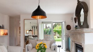 Een woonkamer met een bank, salontafel en een open haard. Er hangt een spiegel aan de muur en een deur leidt naar een tuin.
