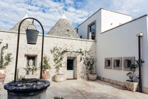 A courtyard with a stone fountain, potted plants, and a building with a white facade and stone roof.
