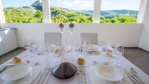 Une table à manger dressée pour six personnes, avec des verres à vin, des assiettes et des couverts, donnant sur un paysage verdoyant grâce à de grandes fenêtres.