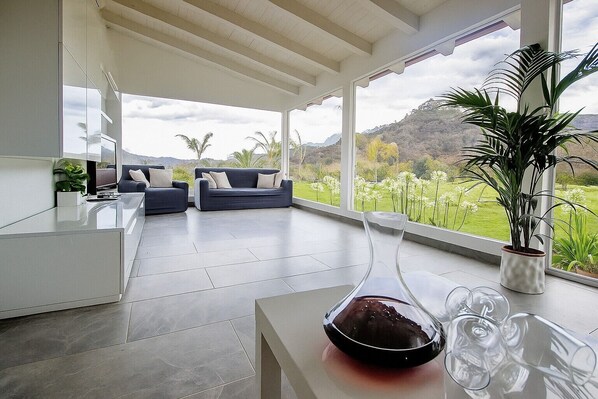 A modern living room with a large window, a sofa, a coffee table, and a potted plant. - Villa 'Eden' with Mountain View, Wi-Fi and Air Conditioning (Castiglione di Sicilia)