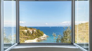 Une vue panoramique depuis une pièce dotée de grandes baies vitrées, donnant sur un paysage côtier caractérisé par un terrain rocheux et une mer d’un bleu limpide.