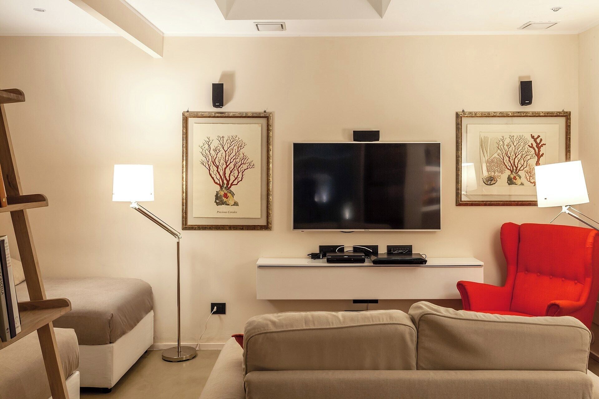 Uma sala de estar moderna com uma TV de tela plana, duas obras de arte emolduradas, uma poltrona vermelha, um sofá bege e um abajur.