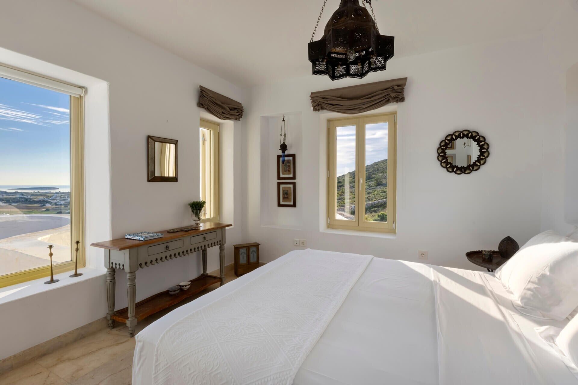 A bedroom with a large bed, a vanity with a mirror, and two windows offering a view of the landscape.