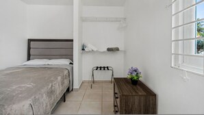 Un dormitorio con cama, mesita de noche, una silla, estante con objetos y ventana con vistas.