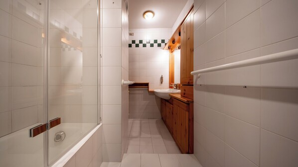 A narrow bathroom with white tiles, a glass shower enclosure, a wooden vanity with a sink, and a checkered tile backsplash.