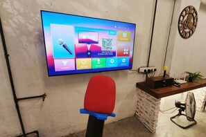 Habitación con una televisión de pantalla plana en la pared, un escritorio con computadora, una silla y un pequeño ventilador.