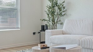 Ett modernt vardagsrum med en vit soffa, ett runt soffbord, en krukväxt och ett fönster med persienner.