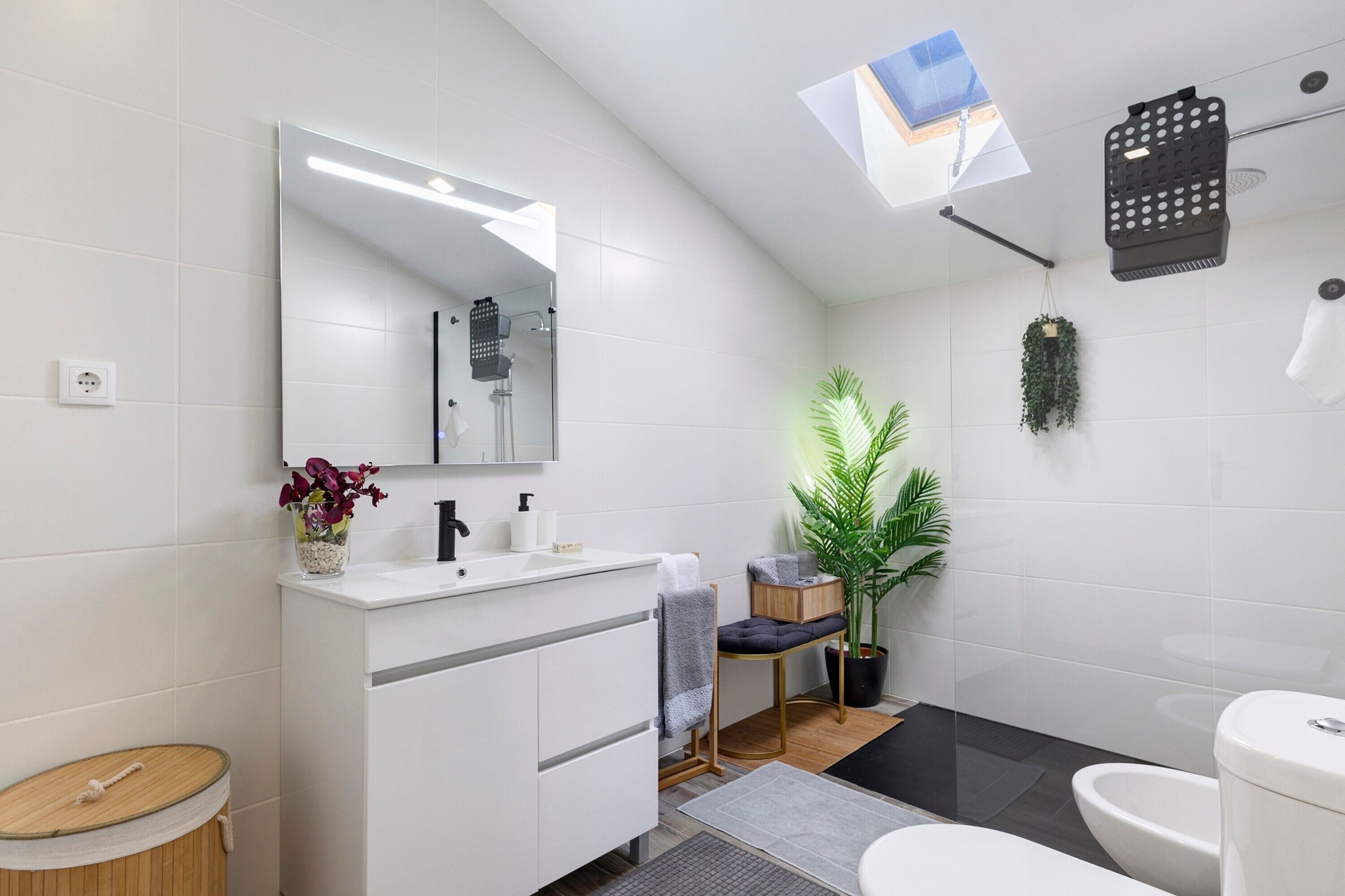 A modern bathroom with a large mirror, white tiles, a wooden floor, a toilet, a bidet, a sink with a towel, and a potted plant.